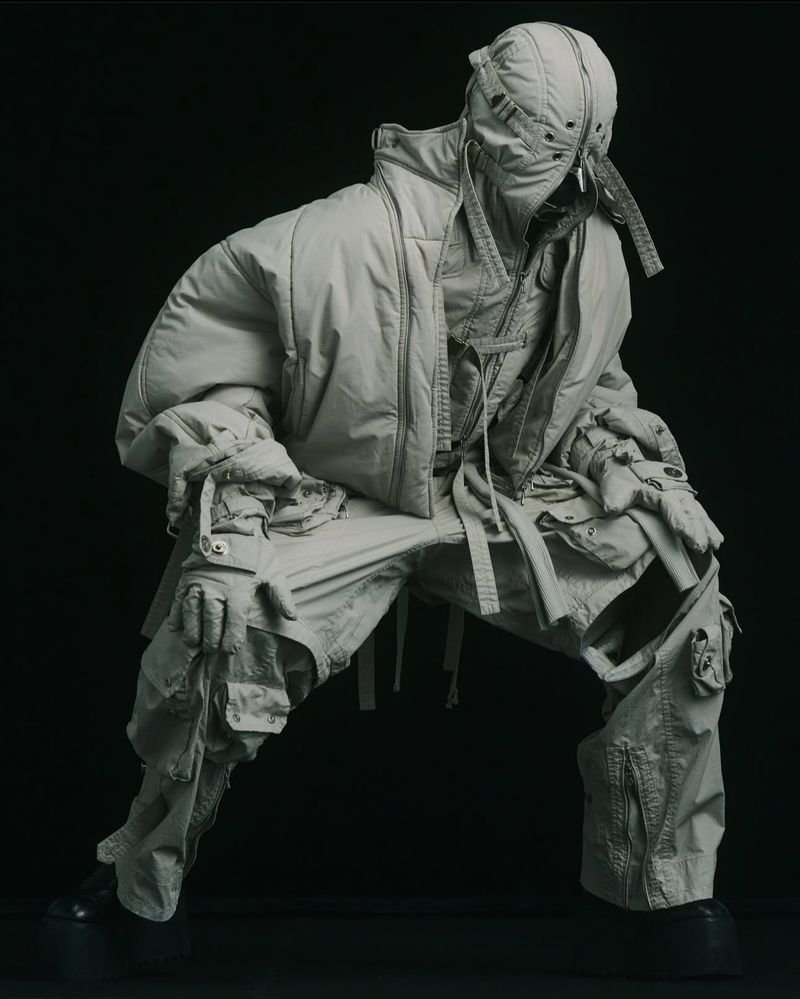 Person in avant-garde fashion, crouching in neutral tones against black backdrop; quilted bomber jacket, balaclava, cargo pants, boots.