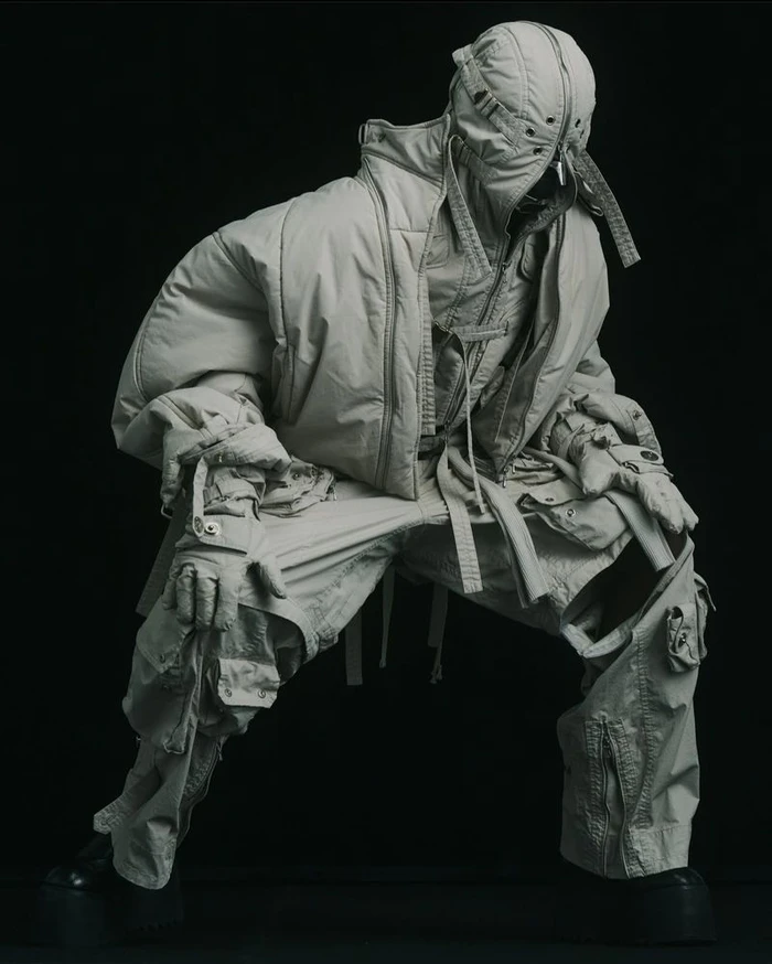 Model wearing a layered hooded grey quilted jacket and matching utility trousers against a solid dark background.