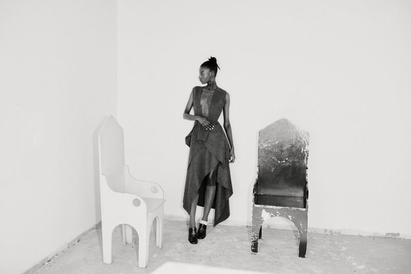 A woman in a dress stands between two art chairs in a white studio, creating a minimalist fashion image with monochrome tones.