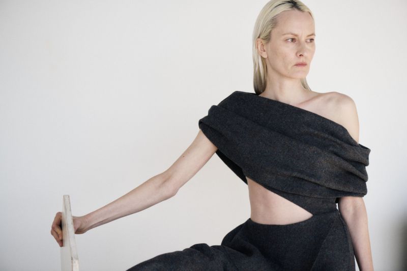 Blonde model in an avant-garde gray two-piece outfit poses in front of a plain white wall in a studio setting.