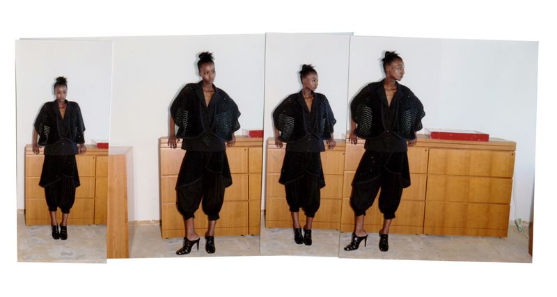 Fashion shot of a black female adult in black culottes, heels, and a cropped jacket with a cabinet in the background.