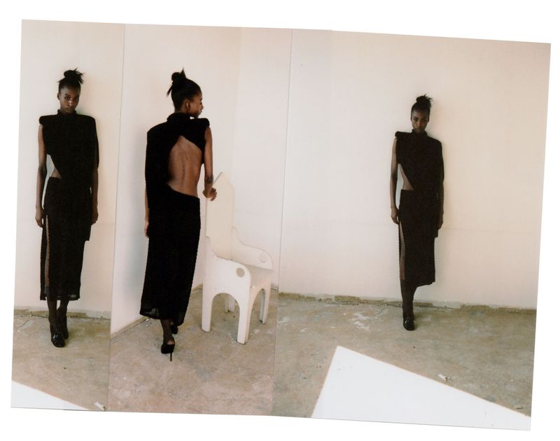 Fashion model wearing a dark, modern dress with an open back poses in front of a white wall in a studio with an abstract white chair.