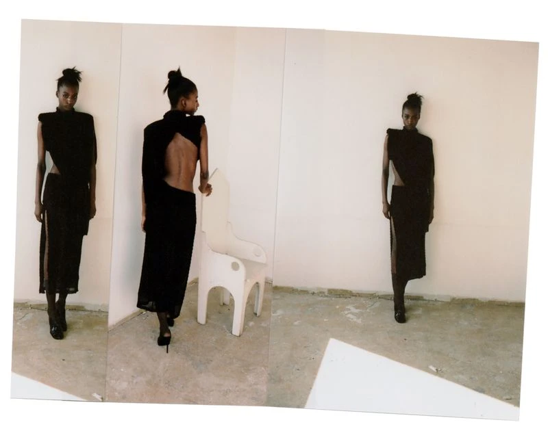Fashion model wearing a dark, modern dress with an open back poses in front of a white wall in a studio with an abstract white chair.