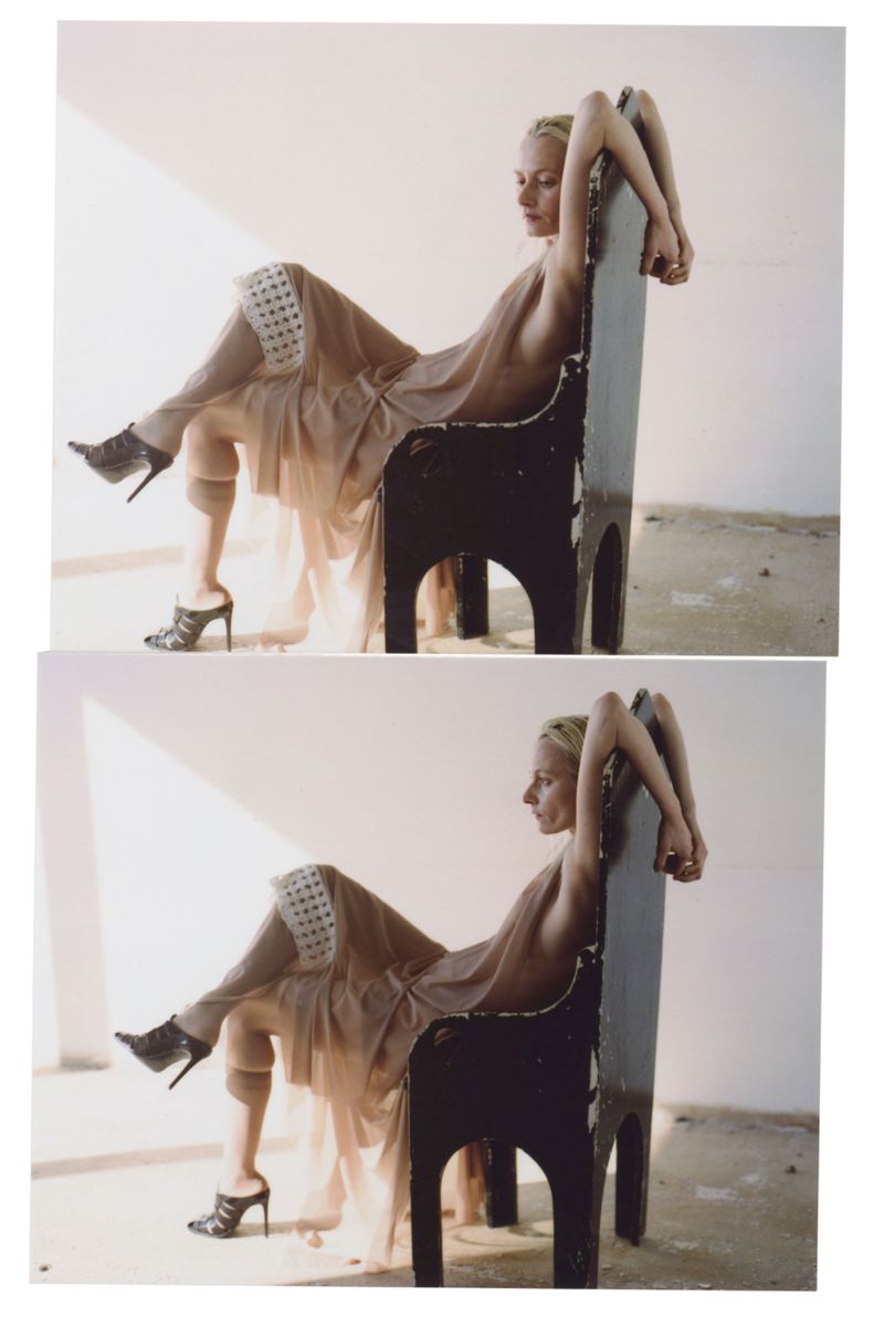 A woman sits backward on a black chair in a photo studio, wearing nude-toned dress and high heels, posing in front of white walls.