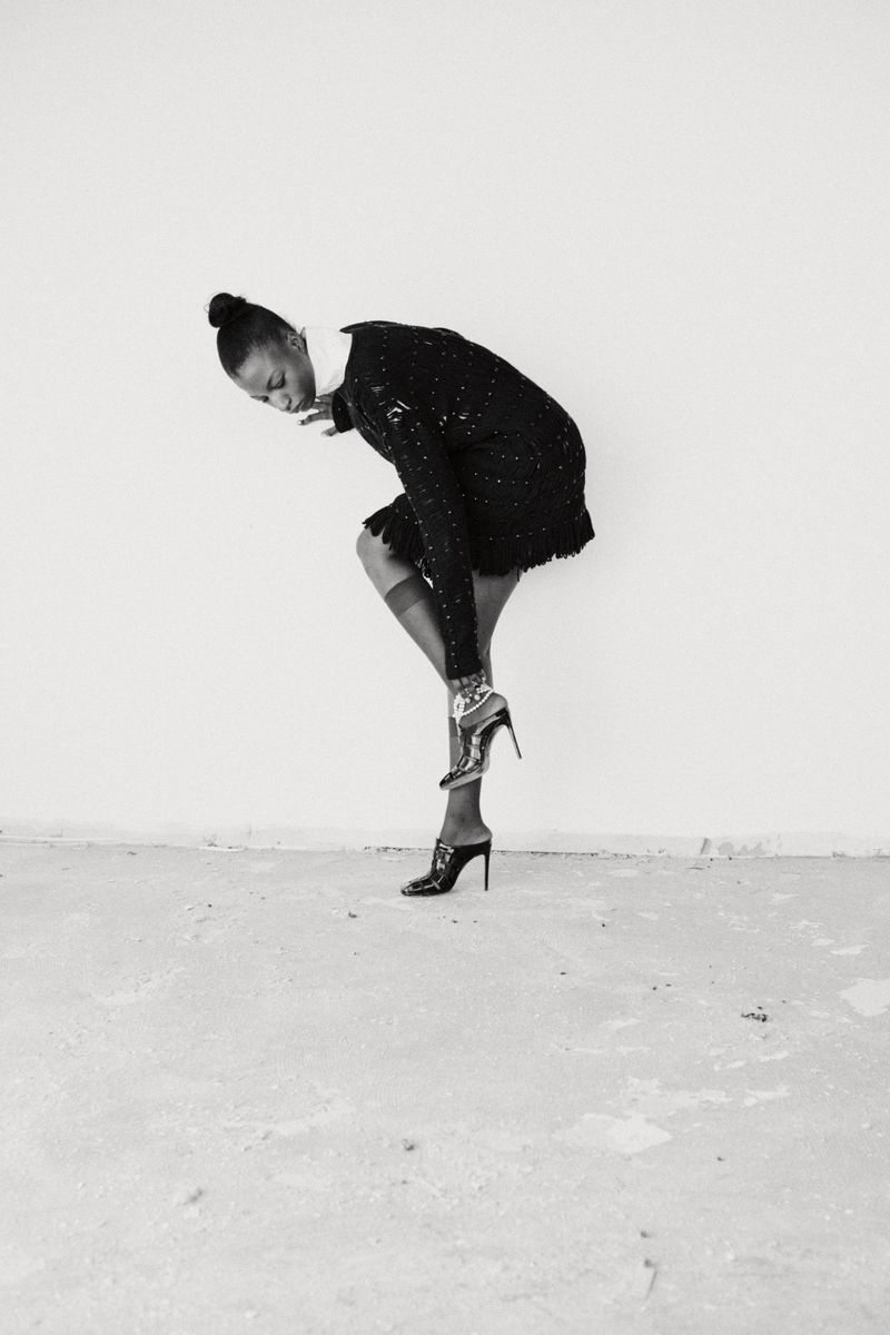 Elegant Black woman in a black sequin dress adjusting her high heel in a minimalist white studio, showing a sophisticated and chic fashion moment.