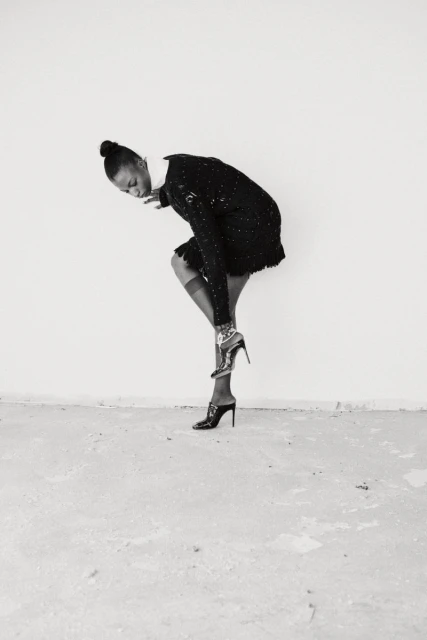 Elegant Black woman in a black sequin dress adjusting her high heel in a minimalist white studio, showing a sophisticated and chic fashion moment.