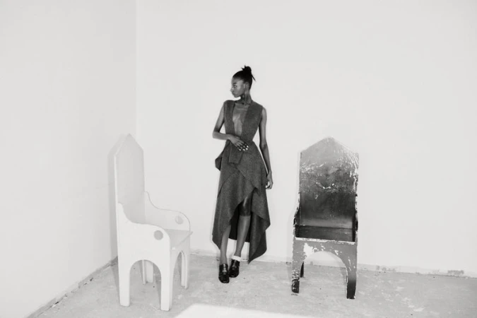 A model standing between two distinctive wooden chairs in a spare room for Diotima Fall/Winter 25.