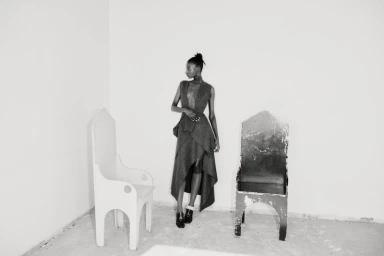 A model standing between two distinctive wooden chairs in a spare room for Diotima Fall/Winter 25.