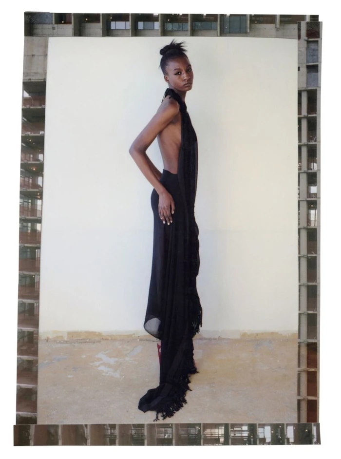 Model in a black backless dress standing against a white backdrop framed by architectural concrete elements