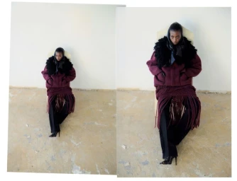Model seated on a chair in a maroon fringed skirt and dark coat against a plain wall.