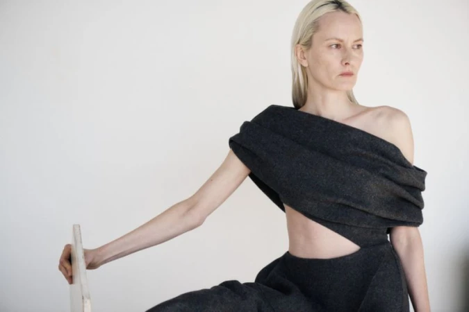 Model with blonde hair wearing dark grey draped sculptural fashion garment against a plain white studio wall.