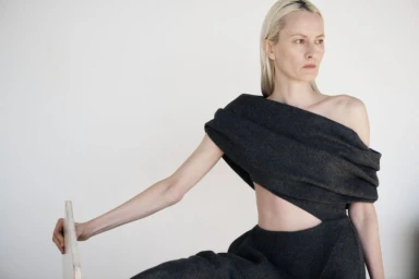 Model with blonde hair wearing dark grey draped sculptural fashion garment against a plain white studio wall.