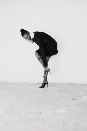 Model in black textured dress and high heels posing against a plain wall in black and white