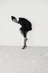 Model in black textured dress and high heels posing against a plain wall in black and white
