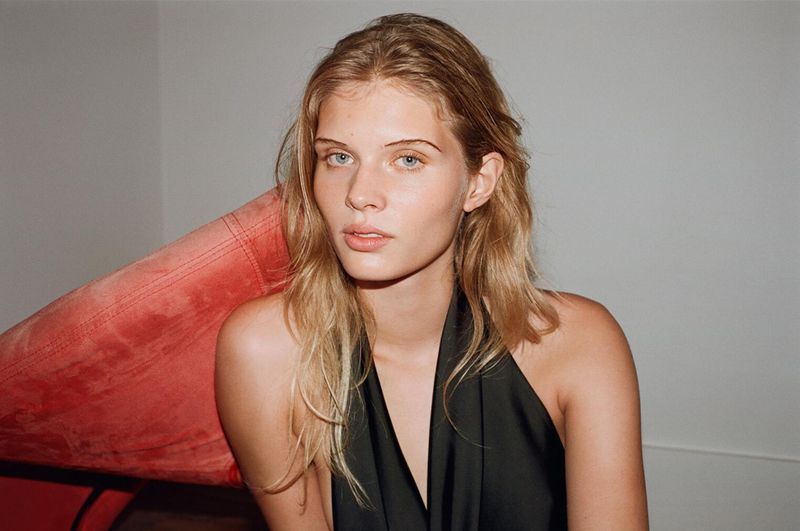 Close-up portrait of a blonde adult female with fair skin posing in a sleeveless black top, indoors, with an orange chair.