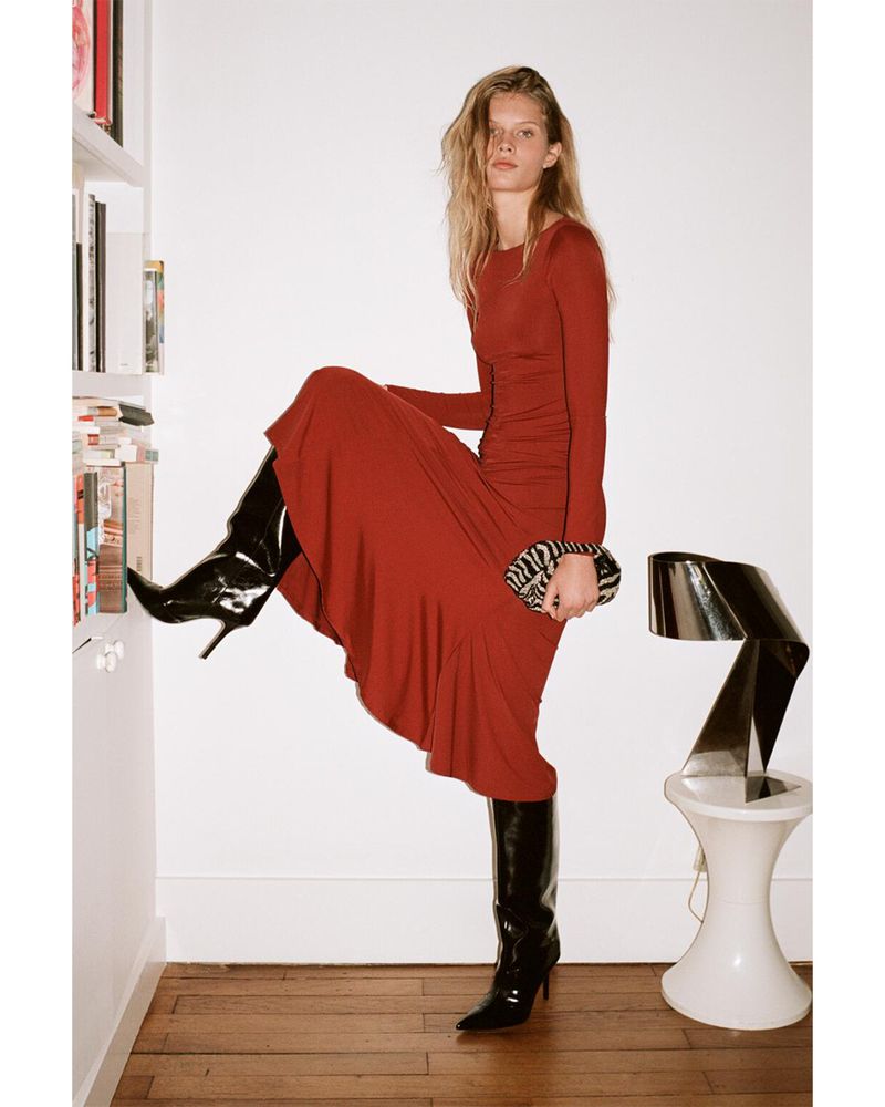 Fashion shot of an adult woman in a red dress and black boots holding a zebra print purse posing next to a bookshelf indoors.