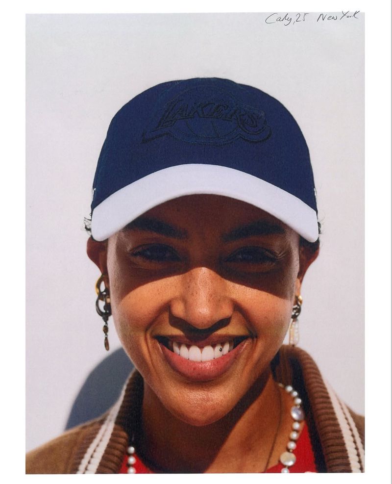 Smiling woman in a dark blue Los Angeles Lakers baseball cap with white brim, wearing gold jewelry and a varsity style jacket.