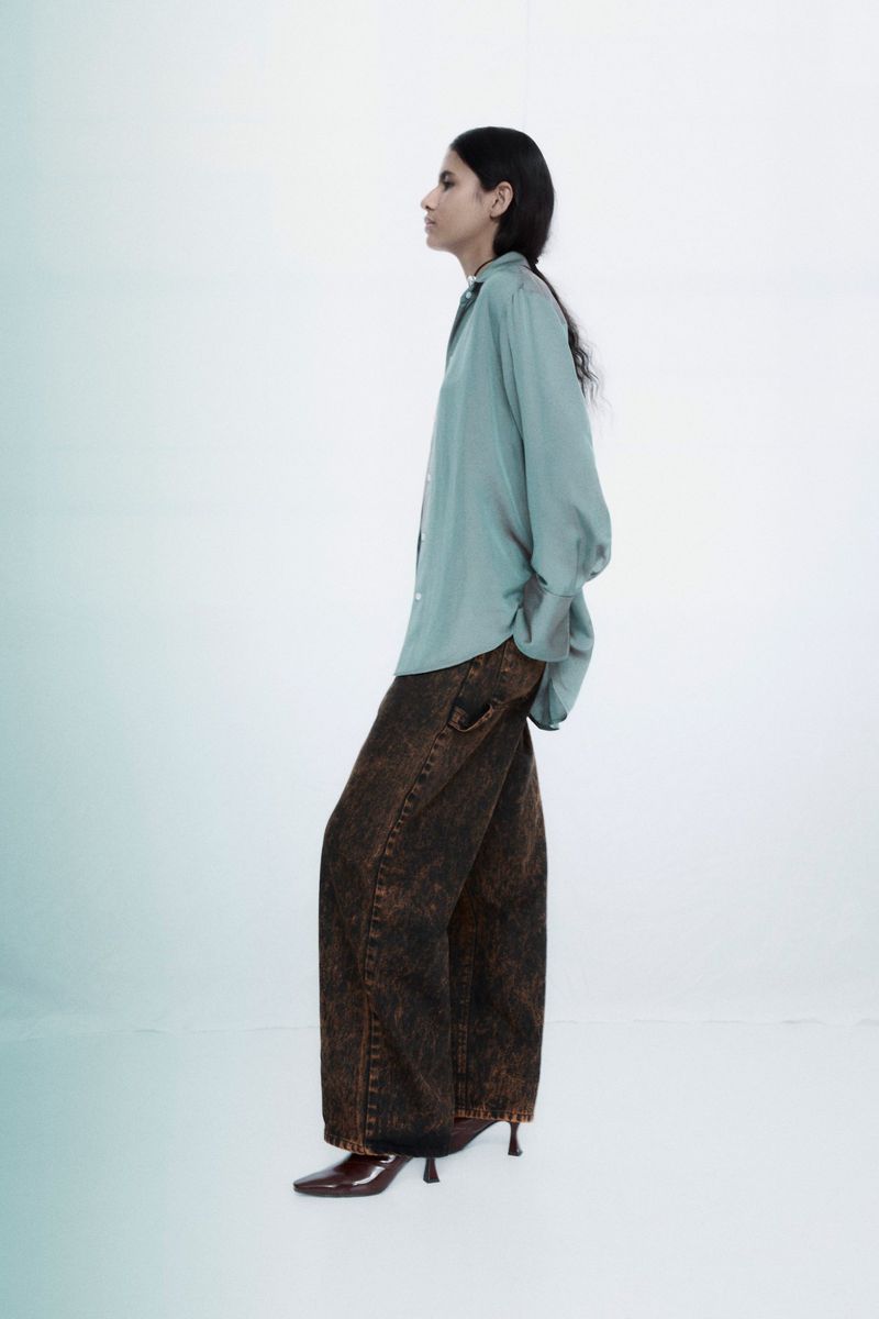 Fashionable adult woman poses in a light blue long-sleeved shirt, brown-toned jeans, and heeled shoes against a gradient background.