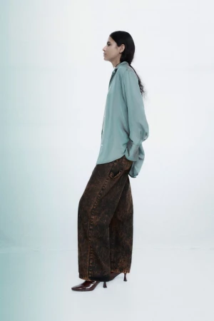 Fashionable adult woman poses in a light blue long-sleeved shirt, brown-toned jeans, and heeled shoes against a gradient background.