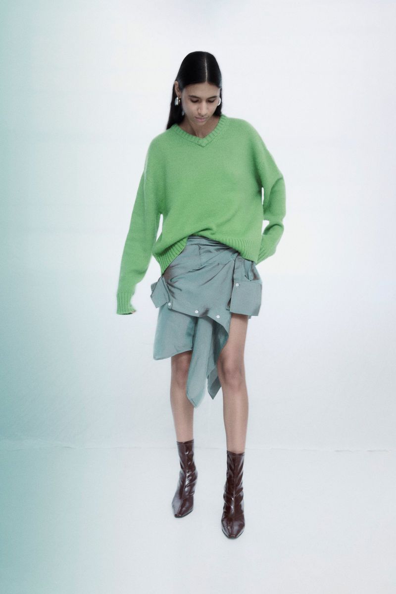 Fashion photo of a woman wearing a green sweater, blue wrap skirt, and brown boots, posing in a photo studio with a white background.