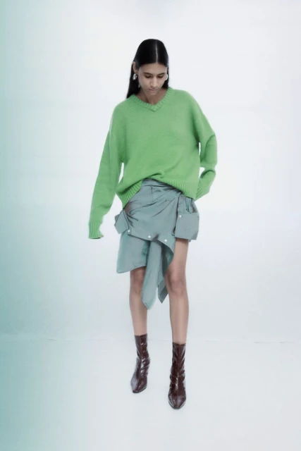 Fashion photo of a woman wearing a green sweater, blue wrap skirt, and brown boots, posing in a photo studio with a white background.