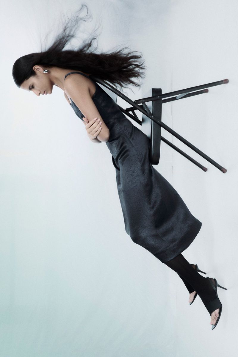 Model wearing a black dress leaning backward against a folding chair against a white background