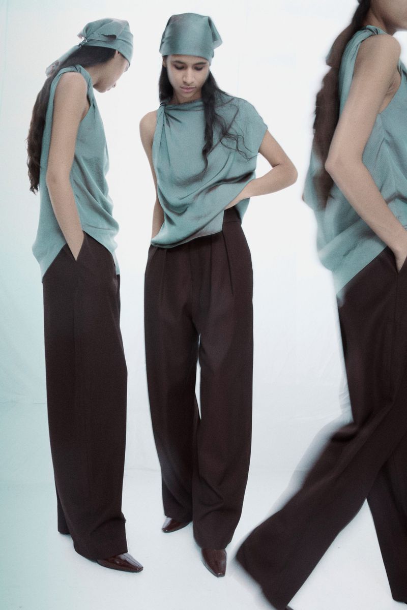 Model Stephanie Quezada wearing a sage green top and dark brown trousers against a white background