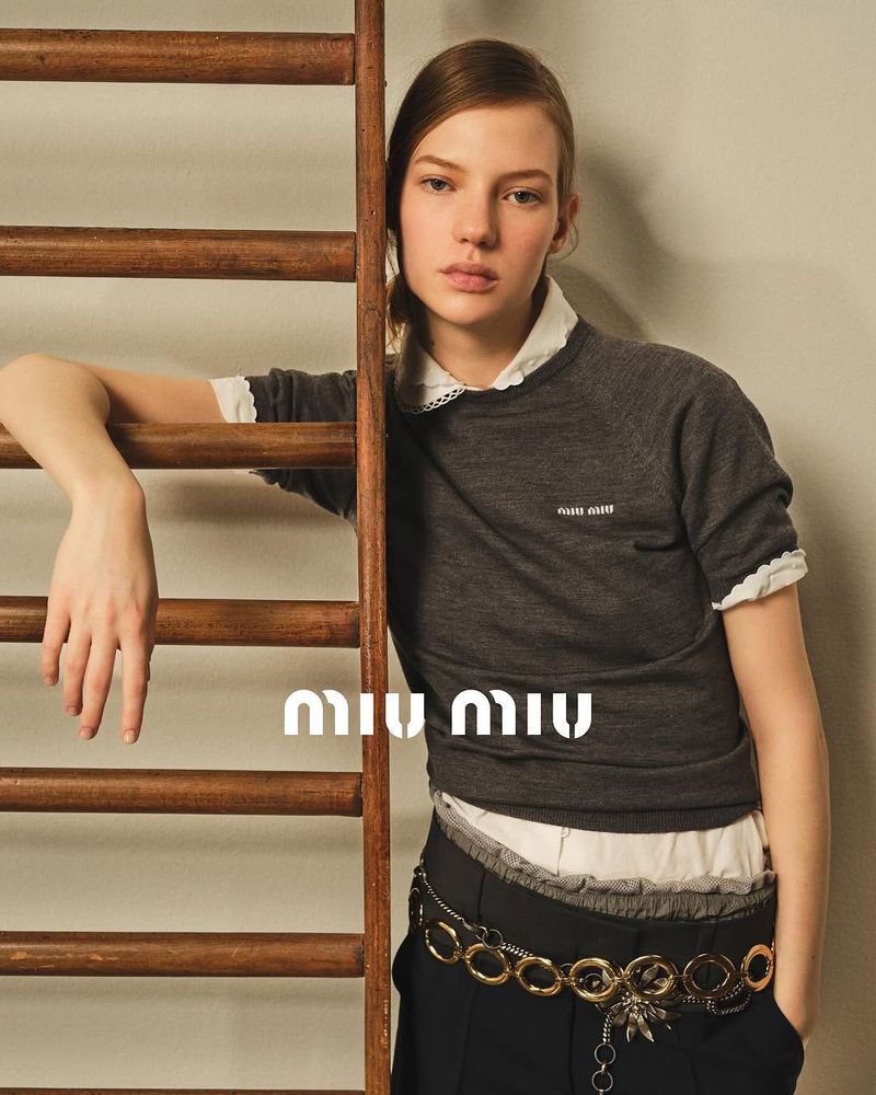 A young woman poses in a fashion shot wearing a grey sweater with a white collar, a gold chain belt, and dark pants next to a wooden ladder.