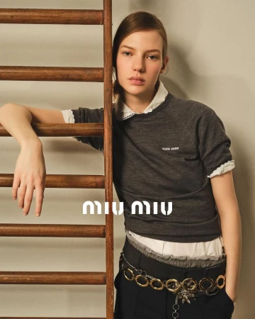 A young woman poses in a fashion shot wearing a grey sweater with a white collar, a gold chain belt, and dark pants next to a wooden ladder.