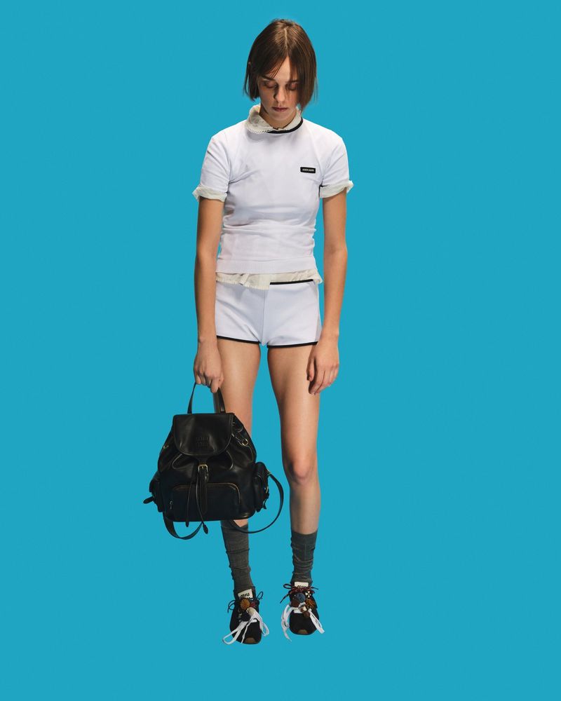 Woman in white top and shorts with black backpack against a blue background in a fashion-forward studio shot, full-length.