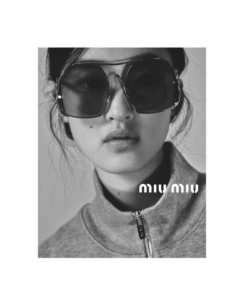 Black and white portrait of a young Asian woman wearing oversized sunglasses and a grey tracksuit top against a neutral background.