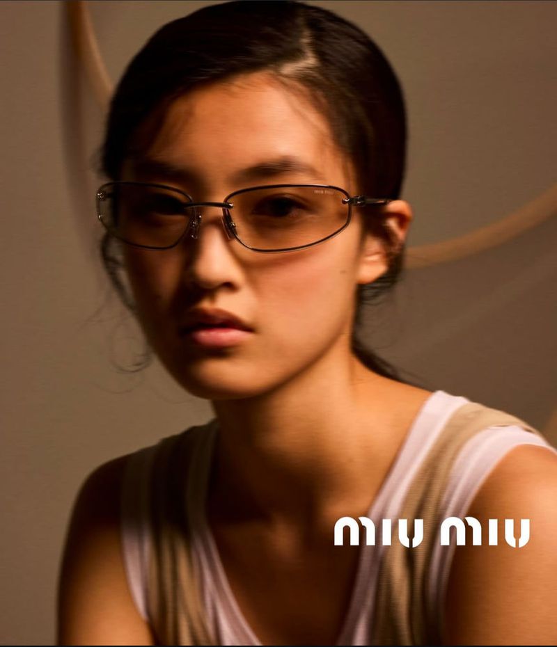 Close-up of an Asian woman wearing glasses and a neutral top against a blurred background, with the Miu Miu logo at the bottom.