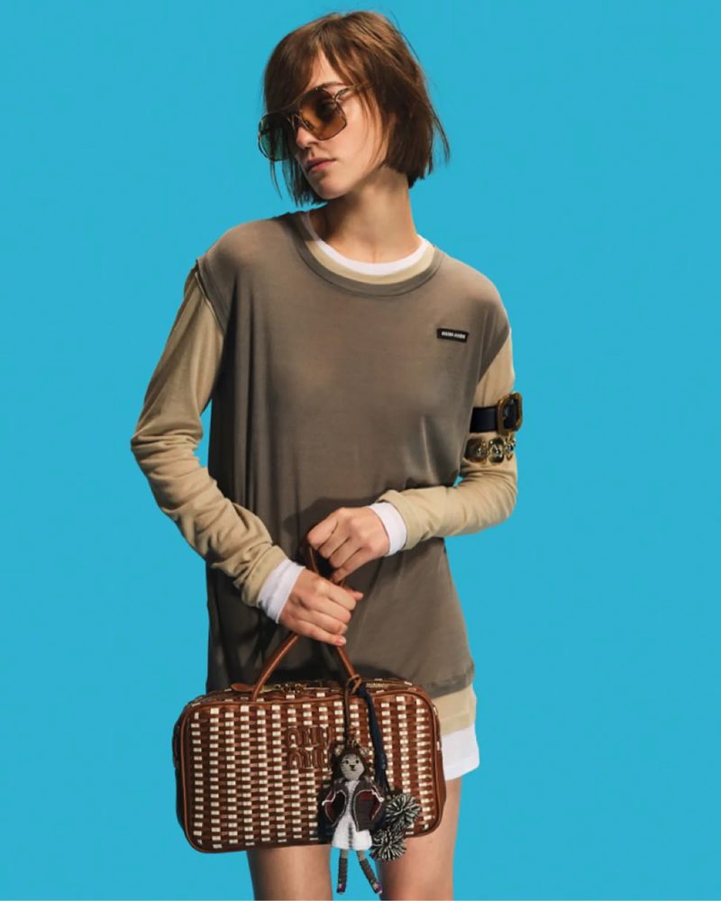 Woman with short brunette hair wears sunglasses and a brown top, holding a woven handbag against a bright blue backdrop.