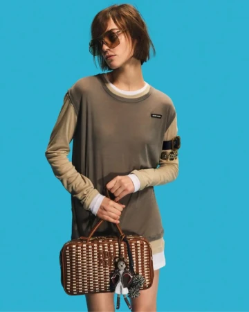 Woman with short brunette hair wears sunglasses and a brown top, holding a woven handbag against a bright blue backdrop.