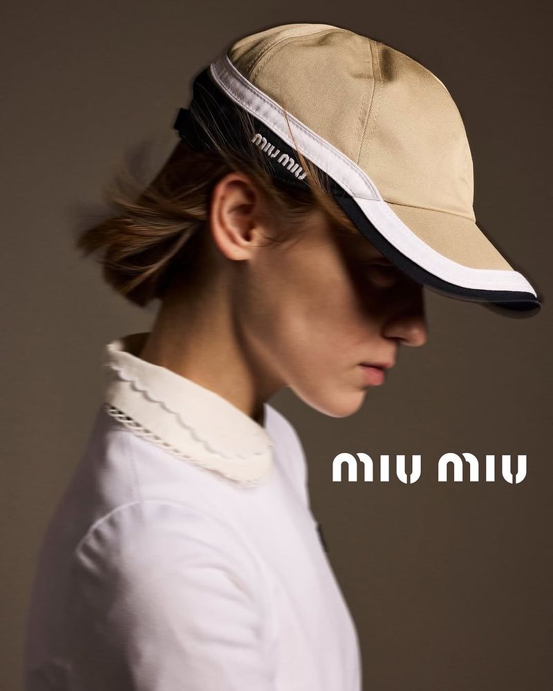 Close-up studio shot of a young adult woman wearing a khaki Miu Miu visor, complemented by a white top with a lace-trimmed collar.
