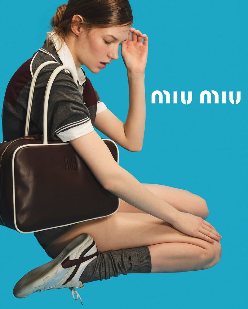 Fashion shot of a young woman sitting with a brown handbag, wearing gray socks and white sneakers against a blue background with a logo.