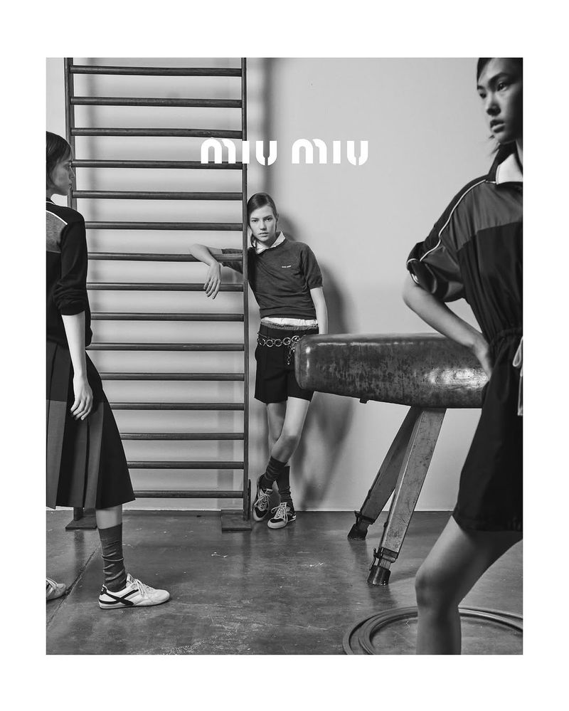 Black and white image of three fashion models in athletic wear, posing in a gymnasium with equipment and the Miu Miu logo.