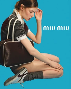 A woman crouching against a solid cyan background holding a dark brown leather shoulder bag, wearing a grey polo shirt and white sneakers.