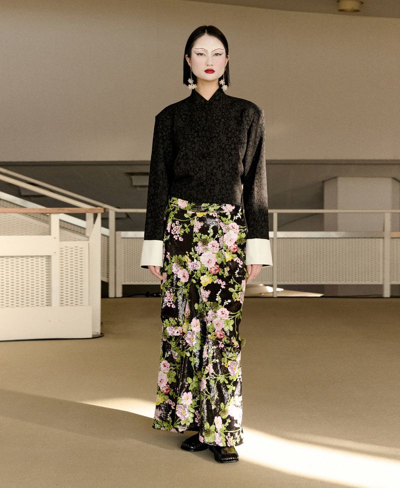 Elegant adult woman standing in an indoor location wearing a black top and floral pants, posing in front of a beige-colored wall.