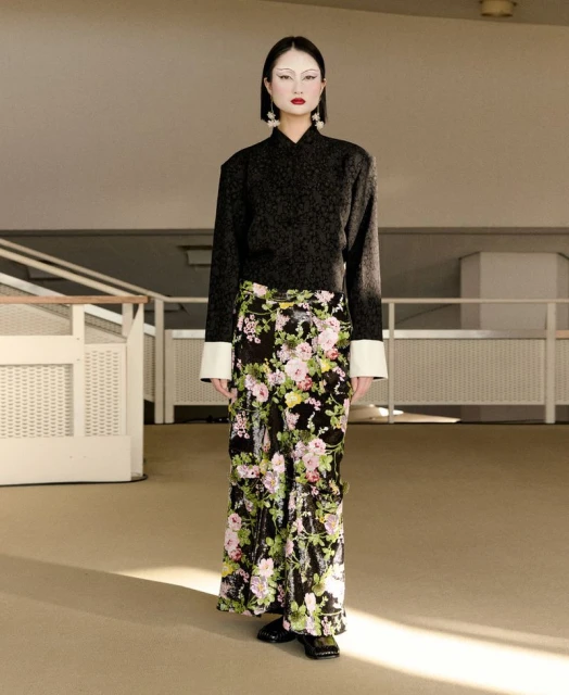 Elegant adult woman standing in an indoor location wearing a black top and floral pants, posing in front of a beige-colored wall.