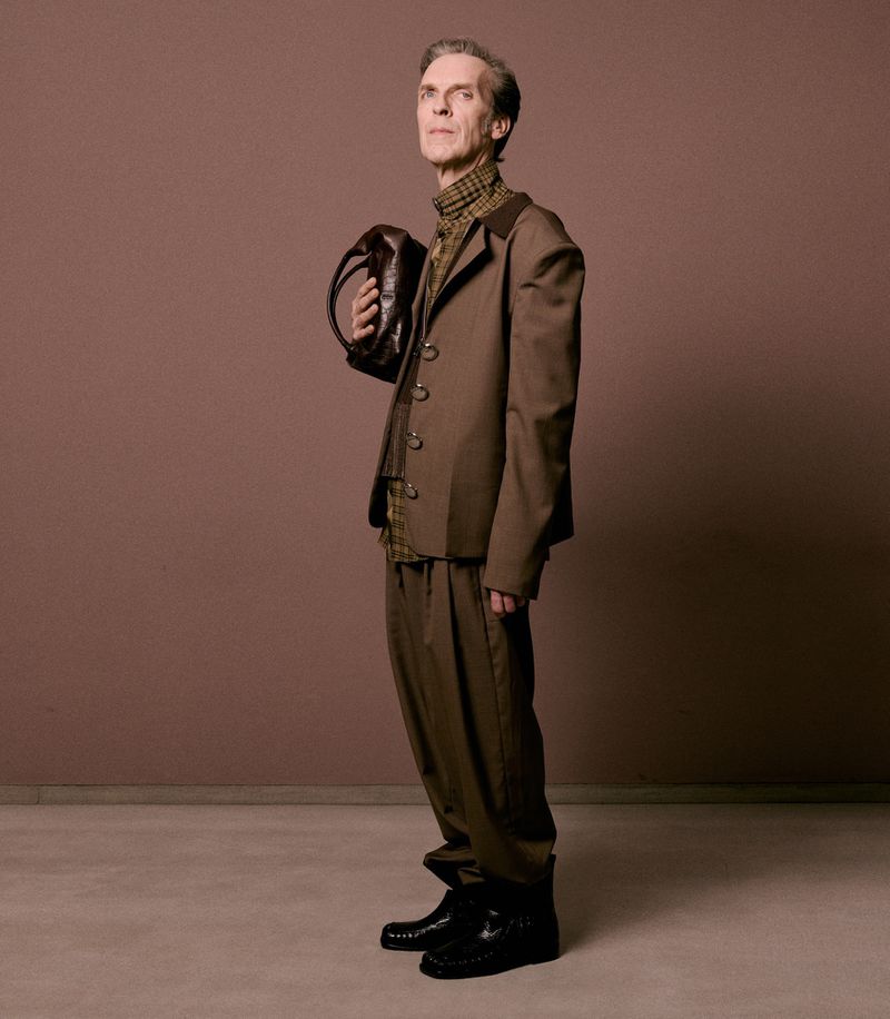 Elderly man in a brown suit holding a purse, standing against a solid brown background in a studio shot.