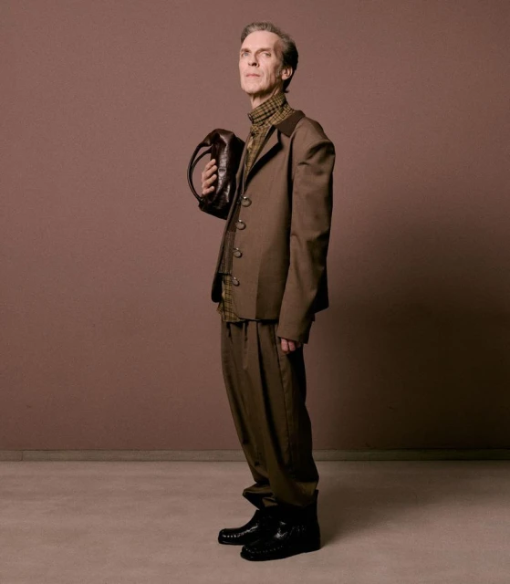 Elderly man in a brown suit holding a purse, standing against a solid brown background in a studio shot.
