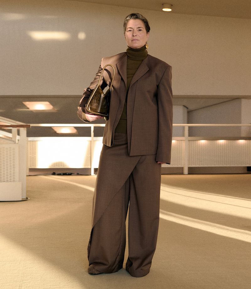 Mature woman in a brown tailored suit standing in a modern indoor space, holding a dark purse, showcasing a professional and stylish fashion look.