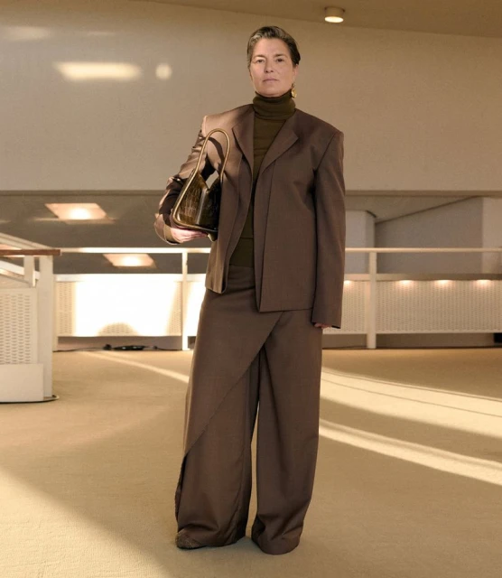 Mature woman in a brown tailored suit standing in a modern indoor space, holding a dark purse, showcasing a professional and stylish fashion look.
