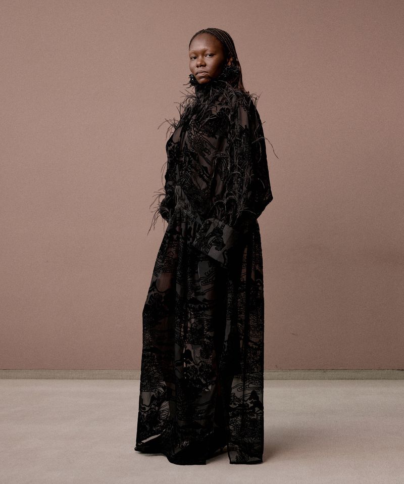 Elegant Black woman in a black feather-trimmed gown poses against a mauve and taupe background in a fashion photography studio.