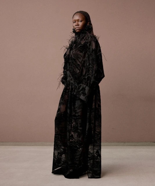 Elegant Black woman in a black feather-trimmed gown poses against a mauve and taupe background in a fashion photography studio.