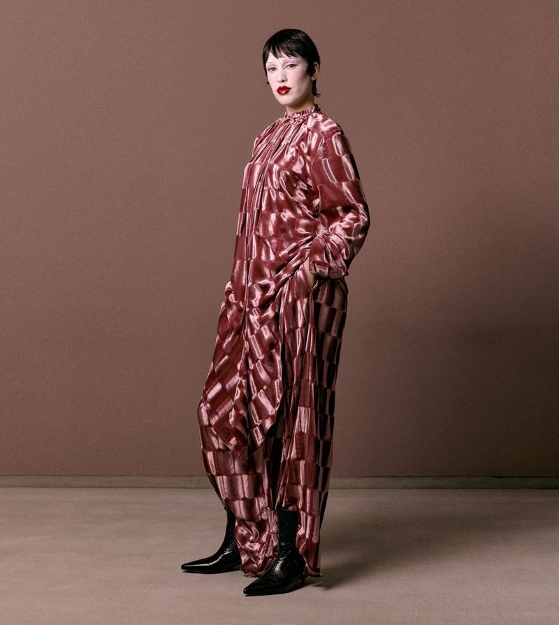 Woman in a brown patterned silk jumpsuit and black boots stands in a photo studio against a brown backdrop. Fashion editorial shot.