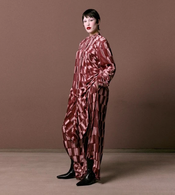 Woman in a brown patterned silk jumpsuit and black boots stands in a photo studio against a brown backdrop. Fashion editorial shot.