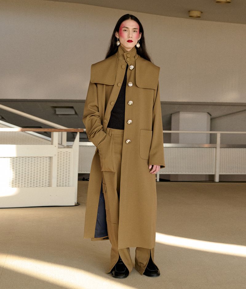 A full-length shot captures a singular adult female showcasing a tan trench coat with statement buttons and matching pants in a neutral setting.