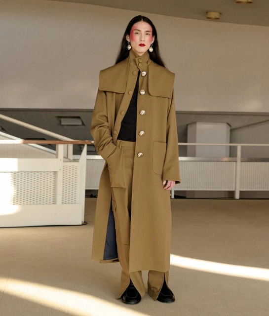 A full-length shot captures a singular adult female showcasing a tan trench coat with statement buttons and matching pants in a neutral setting.